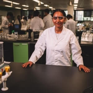 A woman wearing safety glasses and a lab coat stands at a lab bench, with other people working in the background of a laboratory.