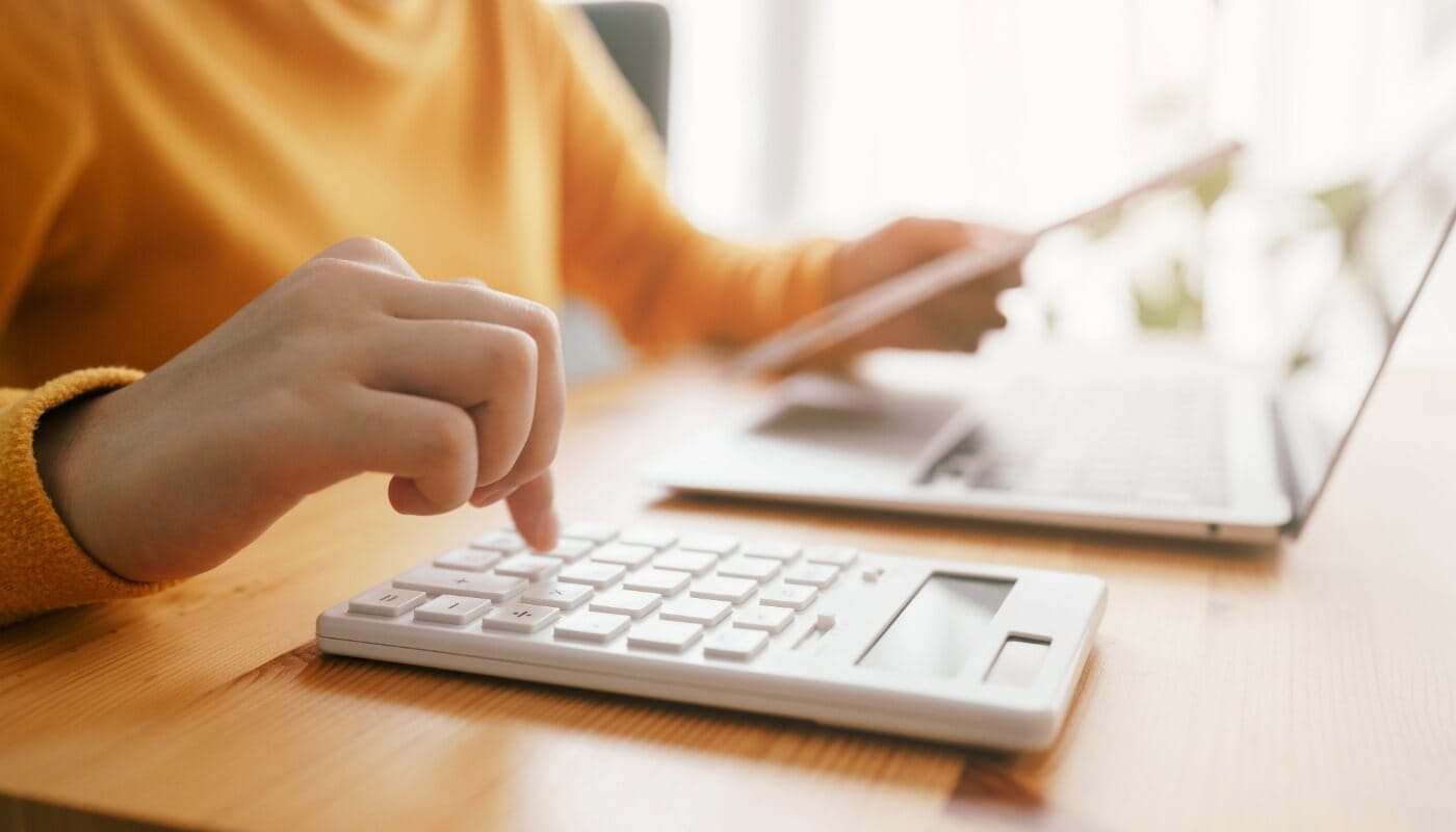 A person uses a white calculator and holds a tablet next to an open laptop on a wooden desk.