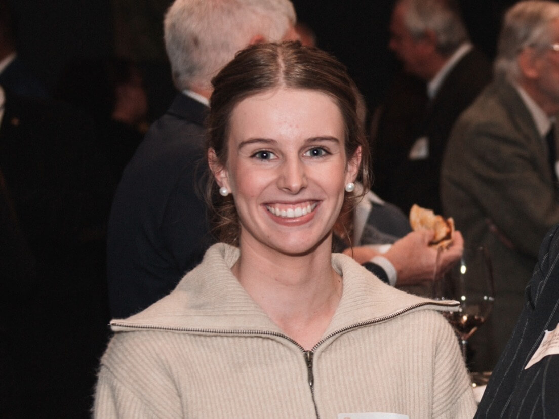 A young woman with brown hair and pearl earrings smiles at an indoor event, surrounded by people in formal attire.