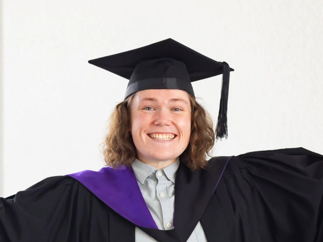 Person smiling broadly while wearing a graduation cap and gown with a purple sash, standing against a plain white background.