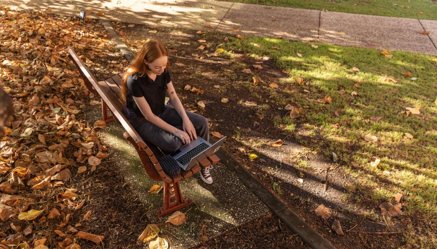 A person with red hair sits on a park bench using a laptop, surrounded by fallen leaves and grass on a sunny day.
