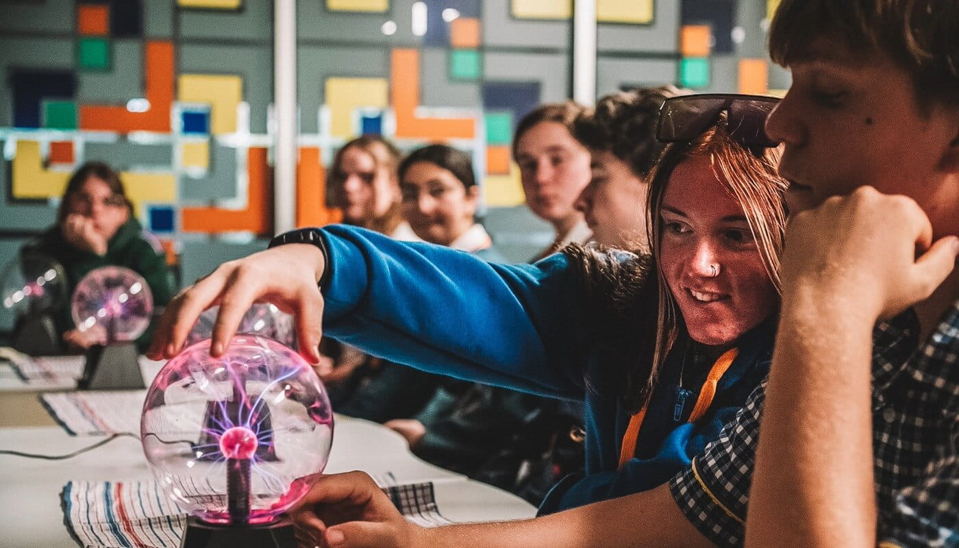 A group of students sits at a table, with one student touching a plasma ball while others watch. Colorful square patterns decorate the wall in the background.