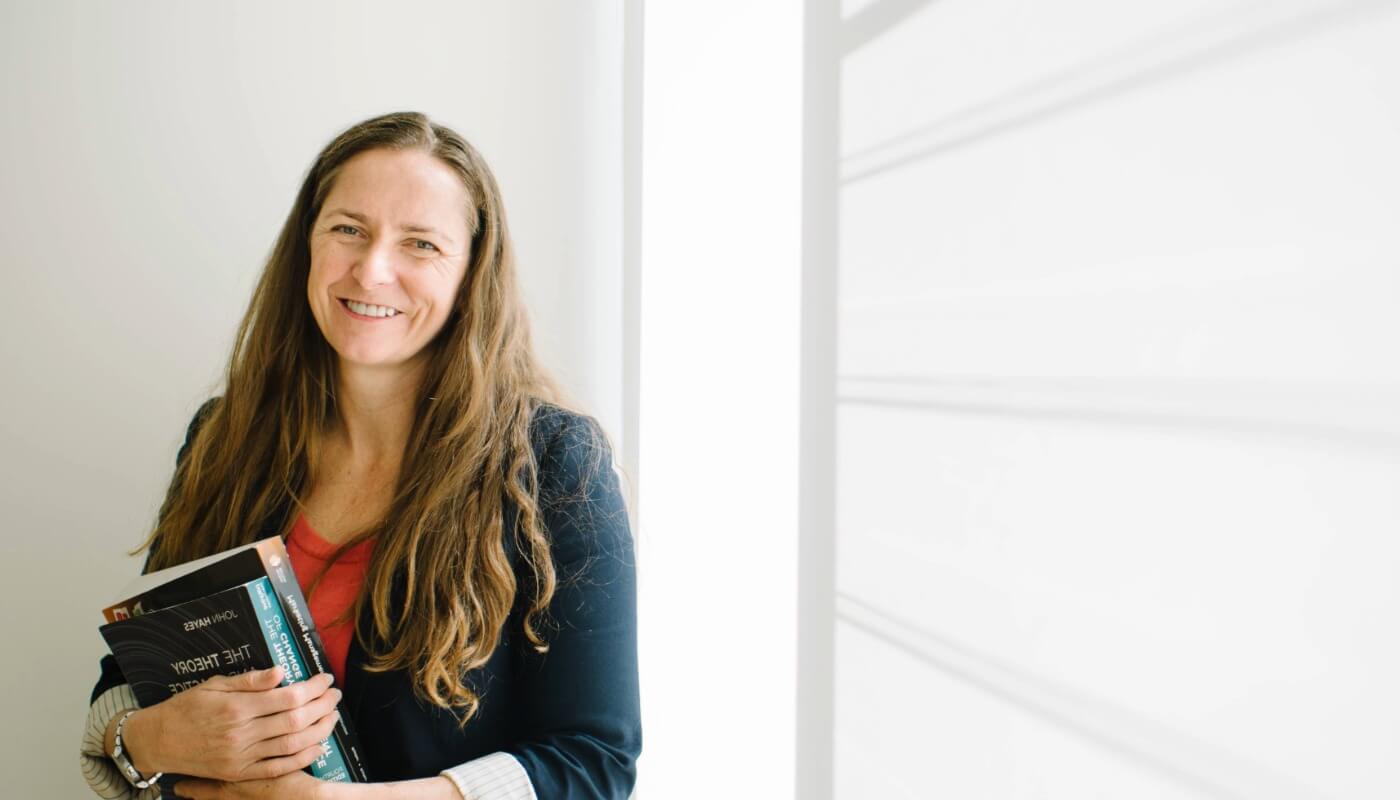 A woman with long brown hair stands by a window, holding books and smiling, wearing a dark blazer over a red shirt.