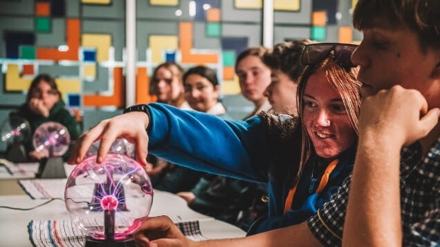 A group of students sit at a table; one student touches a plasma globe, causing pink and purple electric filaments to gather beneath her fingers.