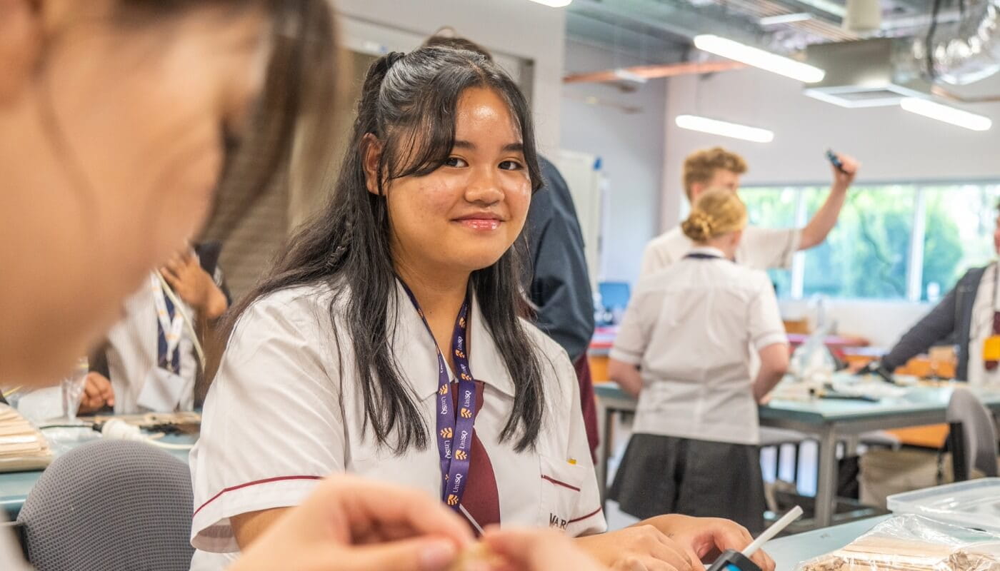 A student in a school uniform sits at a classroom table, smiling at the camera, while other students work and interact in the background.