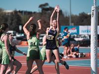 A netball player in a UniSQ uniform jumps to catch the ball near the goal post, while players in green uniforms defend.