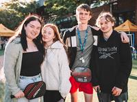 Four young people stand outdoors, smiling at the camera. Two hold pickleball paddles, and tents are visible in the background.