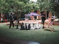 Two people play a large outdoor chess game on artificial grass in a campus courtyard, with a striped deck chair and a canopy tent in the background.