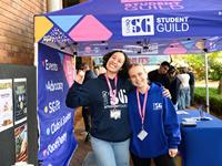Two people in UniSQ Student Guild hoodies stand smiling under a Student Guild tent at an outdoor event, with informational materials on tables nearby.