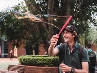 A person wearing a backpack and cap smiles while waving a bubble wand, creating a long soap bubble in an outdoor park area.