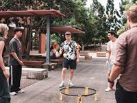 Five young adults play Spikeball outdoors on a paved area, surrounded by trees and benches, with one person about to hit the ball.