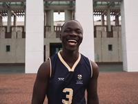 A person wearing a UniSQ basketball jersey with the number 3 is smiling and standing outdoors in front of a large, white building.