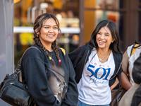 Two young people stand outdoors smiling and talking.