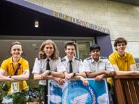 Five young people, three in UniSQ pilot uniforms and two in gold shirts, stand in front of a building labeled "Flight Simulator".