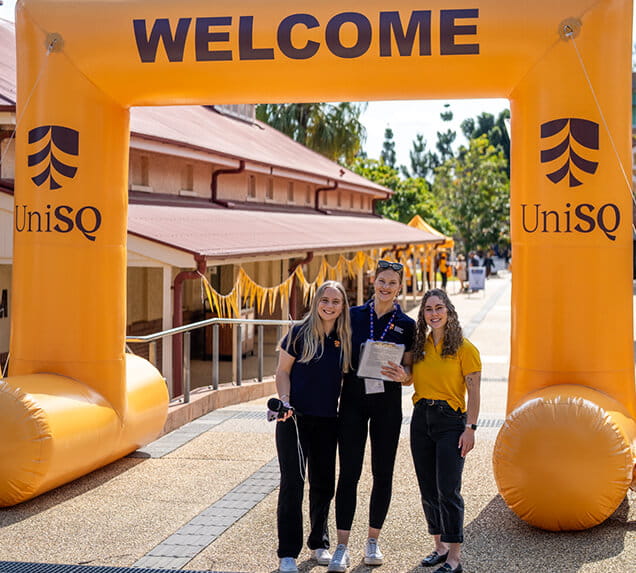 Three people stand and smile under a large yellow "WELCOME" archway displaying the UniSQ logo, with buildings and trees in the background.