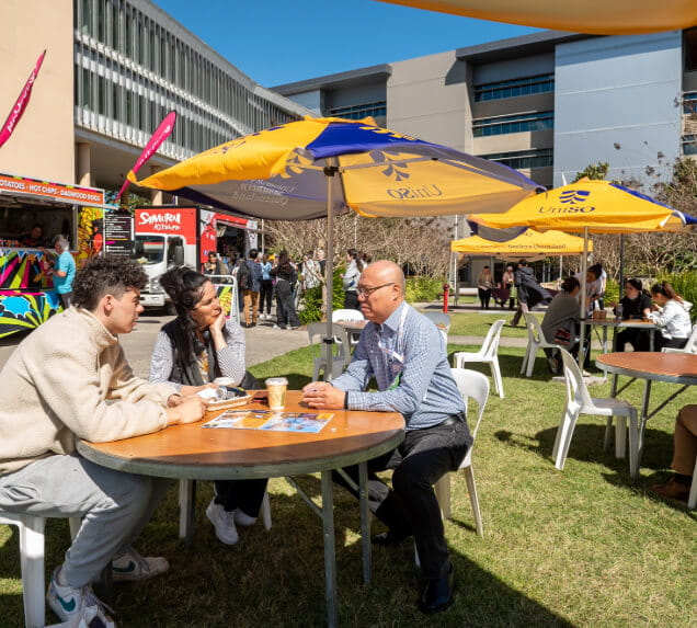 Three people sit at a round outdoor table under an umbrella, talking, with food trucks and other people in the background on a sunny day.