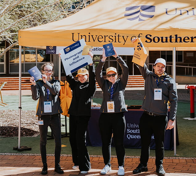 Four people stand in front of a University of Southern Queensland tent holding flags, signs, and lanyards, posing for a photo during an outdoor event.