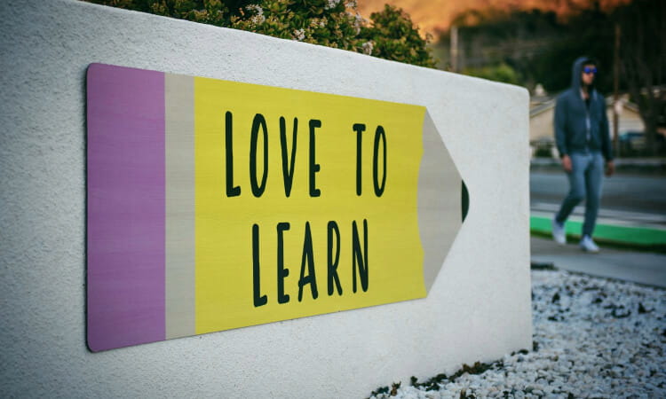 Sign shaped like a pencil with the words "Love to Learn," mounted on a low white wall; a person walks by in the background.