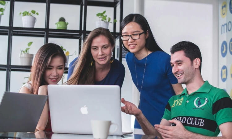 A group of four people gathers around a laptop, engaged in discussion. There are plants on shelves in the background.