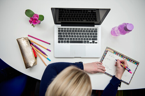 Person at a desk writing in notepad with open laptop and stationery