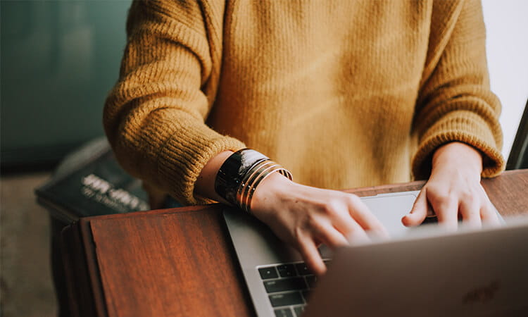 Person in a yellow sweater typing on a laptop with bracelets visible on the wrist.
