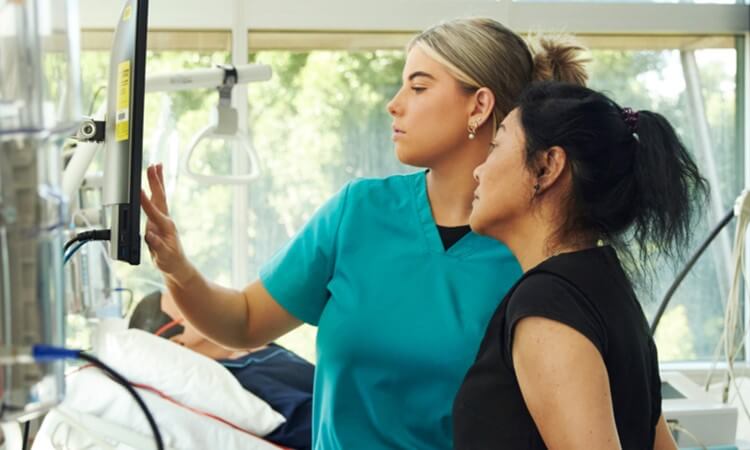 Two healthcare professionals examining a monitor in a medical setting, with a patient lying in the background.