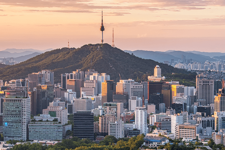 A cityscape view of Seoul with tall buildings in the foreground and Namsan Mountain with Namsan Tower in the background at sunset.