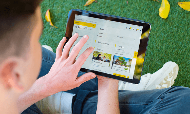 A person using a tablet outdoors, viewing a website or app interface with menu options including 'Student Centre' and 'Library.' Bright grass and scattered leaves surround them.