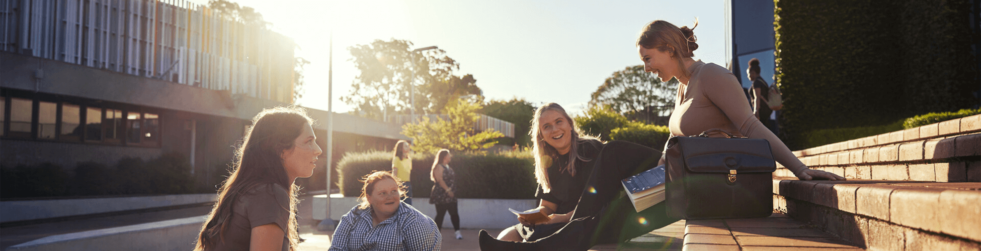 A group of four people sitting outdoors on steps, laughing and talking under the sun. Buildings and trees are in the background.