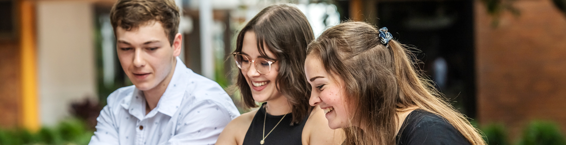 Three young adults sit together outdoors, smiling and looking at something out of view.