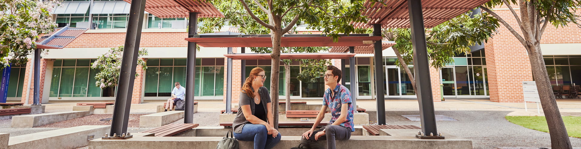 Two people sit facing each other and talking on benches under a shaded outdoor area, with a person sitting in the background near a modern building.