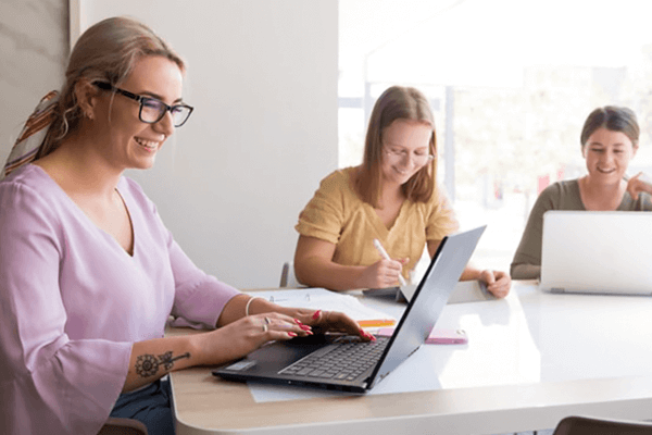 Three people sit at a table, using laptops and taking notes, in a bright room.