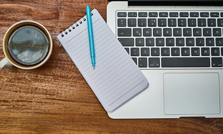 A laptop with a notepad and pen on its keyboard next to a cup of coffee on a wooden desk.