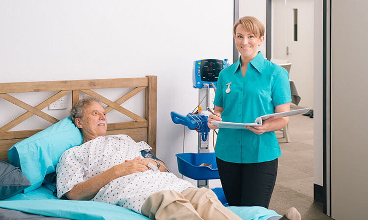 A nurse holding a clipboard stands beside a patient's bed in a hospital room.