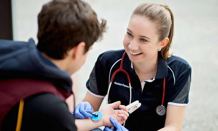 A smiling female healthcare student in a uniform is taking a patient's temperature with a digital thermometer.