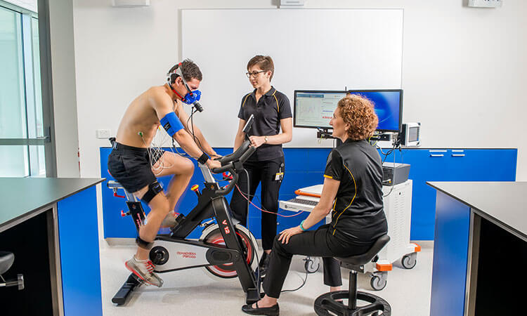 A man undergoes a fitness assessment on a stationary bike while being monitored by two professionals in a clinical setting.