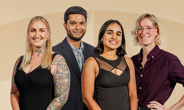 Four people stand side by side, facing the camera and smiling. They are dressed in semi-formal attire against a beige background.