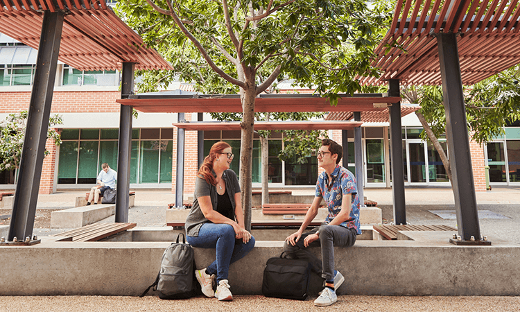 Two people sit and chat on a bench in an outdoor courtyard area at the Ipswich campus, while another person sits on a separate bench in the background. Each has a backpack nearby.