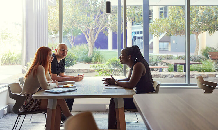 Three people are seated at a table in a bright room with large windows, engaged in a discussion.