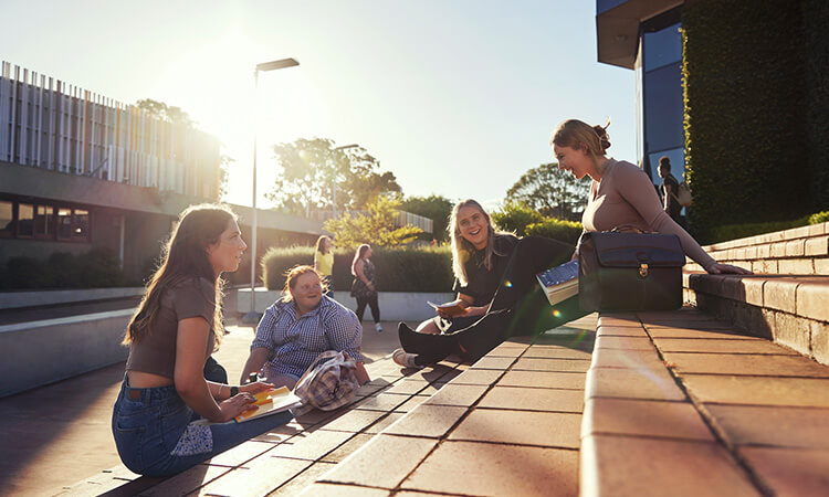 A group of four people sit outdoors on brick steps, engaging in conversation and laughter with a building in the background during daylight.