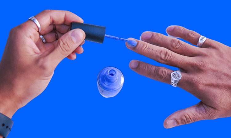 A person holding a nail polish brush in one hand and applying light blue nail polish to one fingernail on the other hand, against a blue background. A bottle of nail polish is open below the hands.