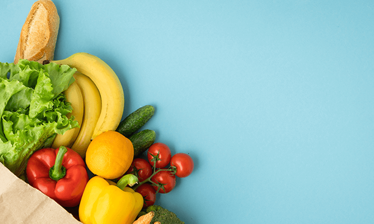 A grocery bag filled with various fresh produce including lettuce, bananas, cucumbers, tomatoes, lemons, peppers, and a baguette against a light blue background.