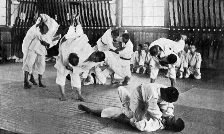 Several pairs of people in martial arts uniforms practice judo techniques in a dojo; some observe while sitting against the wall.