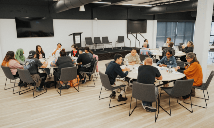 A group of people sits around three round tables in a modern conference room, participating in a discussion or meeting.
