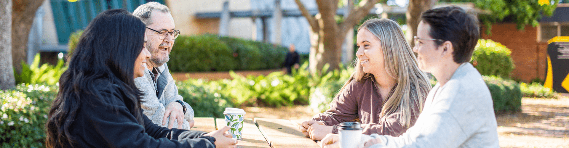 Four people sit at a wooden picnic table outdoors, drinking from mugs and conversing, with trees and a building in the background.