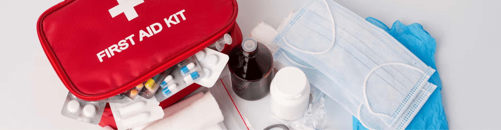 A red first aid kit with medical supplies including pills, a bottle, a thermometer, face masks, gloves, and bandages arranged on a white surface.
