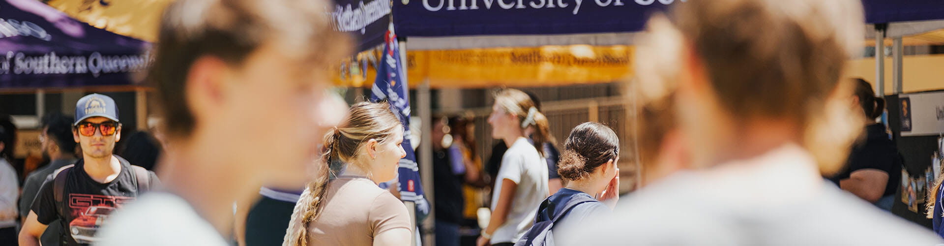Students walk among stalls at an outdoor university event, with banners and tents visible in the background.