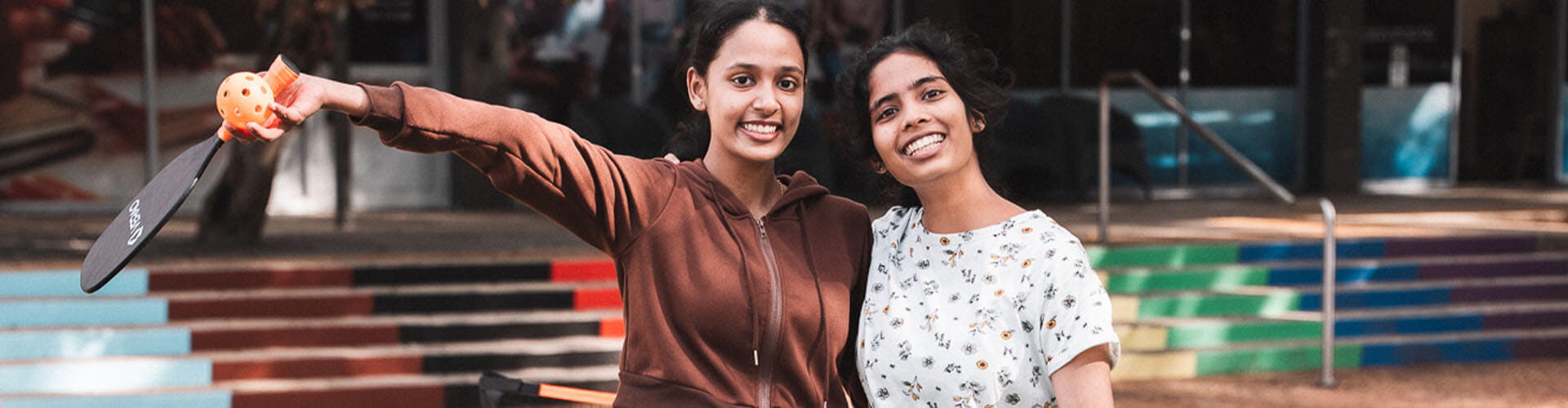 Two young women stand outdoors smiling; one holds a paddle and a ball. Colorful striped steps and a building entrance are visible in the background.
