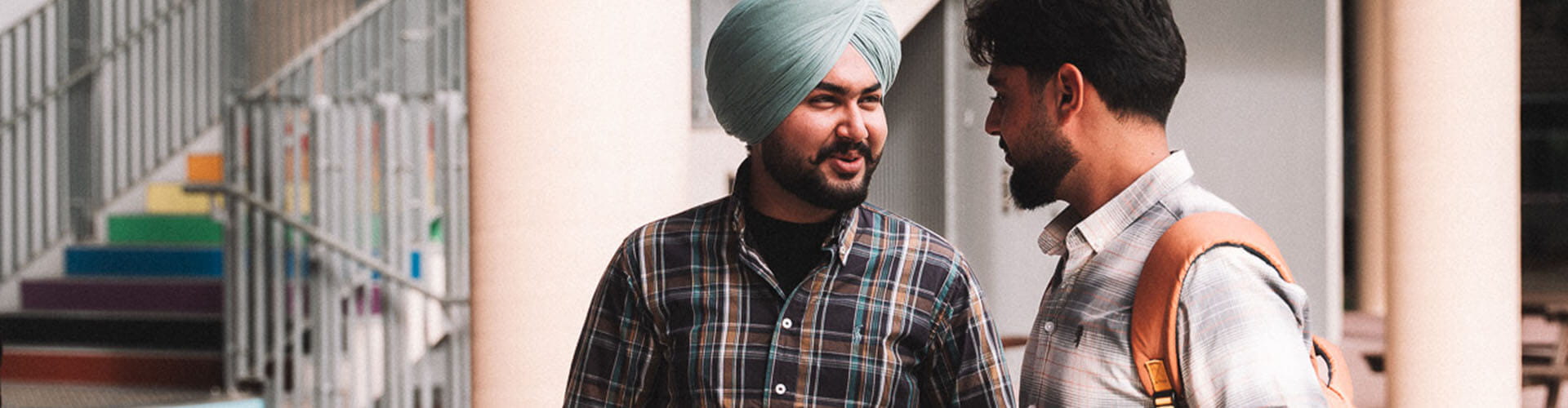 Two men stand and talk outside near a staircase with rainbow-colored steps. One man wears a turban and both are dressed casually.