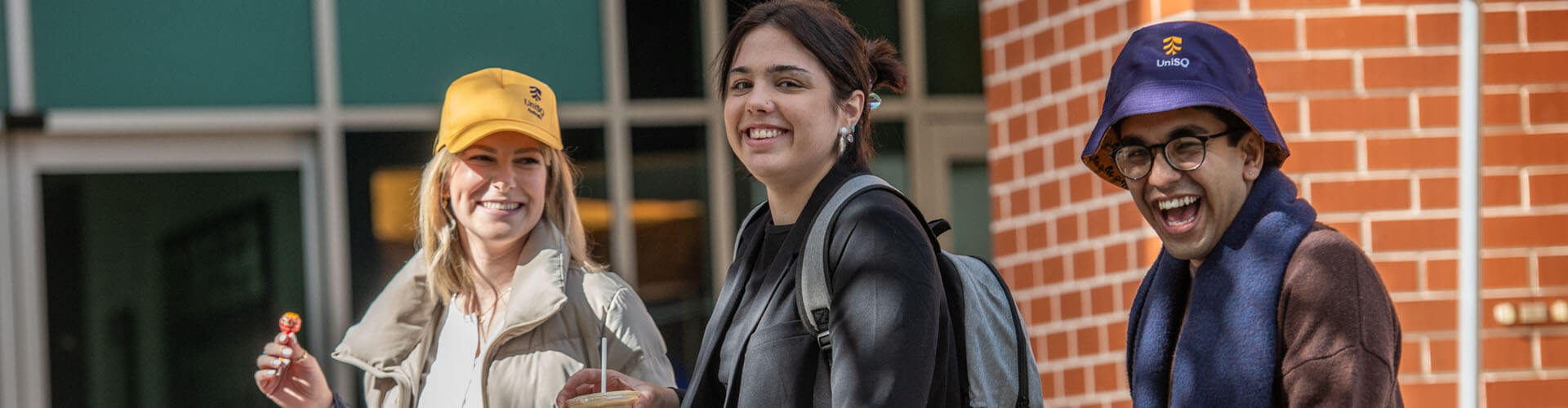 Three people wearing jackets and hats smile while walking outside near a brick wall, one holding a drink and another holding a snack.
